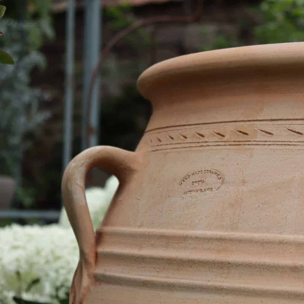 Eine große, hellbraune XXL-Terrakotta-Amphore „Thymus“ mit 100 cm Höhe, geschwungenem Henkel und eingravierten Zierlinien steht im Freien. Auf der Seite des Topfes befindet sich ein Stempel mit schwachem Text. Der Hintergrund zeigt üppiges grünes Laub und eine verschwommene Außenkulisse.