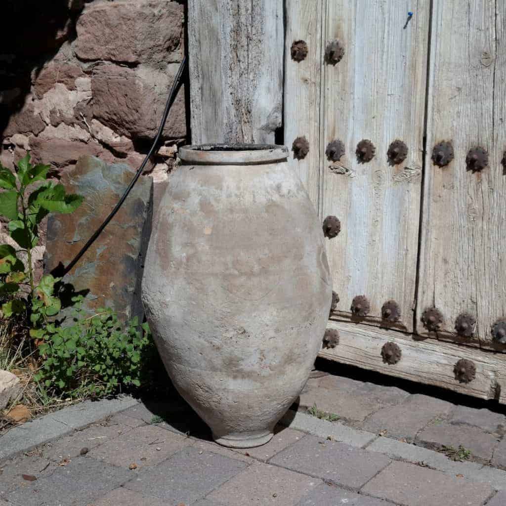 Ein großer, verwitterter Terrakotta-Topf steht auf einer Pflastersteinfläche neben einer groben Holztür, die mit verrosteten Metallknöpfen verziert ist. Neben dem Topf wächst eine kleine grüne Pflanze, und im Hintergrund ist eine Steinmauer zu sehen, die teilweise mit Kletterpflanzen bedeckt ist, was an eine Szene aus einer alten Galerie erinnert.