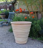 Der winterfester großer Pflanzkübel „Acanthus“, H=60 cm, mit wellenförmigem Linienmuster steht auf einer Kiesfläche in einem Garten. Dahinter blühen rot-orange Mohnblumen, grünes Laub und Holzstühle. Im Hintergrund sind ein Steinpflanzkübel und eine Hauswand zu sehen.