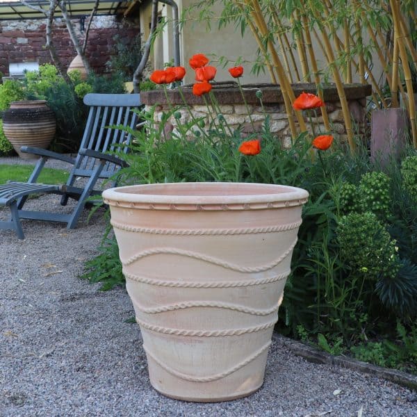 Der winterfester großer Pflanzkübel „Acanthus“, H=60 cm, mit wellenförmigem Linienmuster steht auf einer Kiesfläche in einem Garten. Dahinter blühen rot-orange Mohnblumen, grünes Laub und Holzstühle. Im Hintergrund sind ein Steinpflanzkübel und eine Hauswand zu sehen.