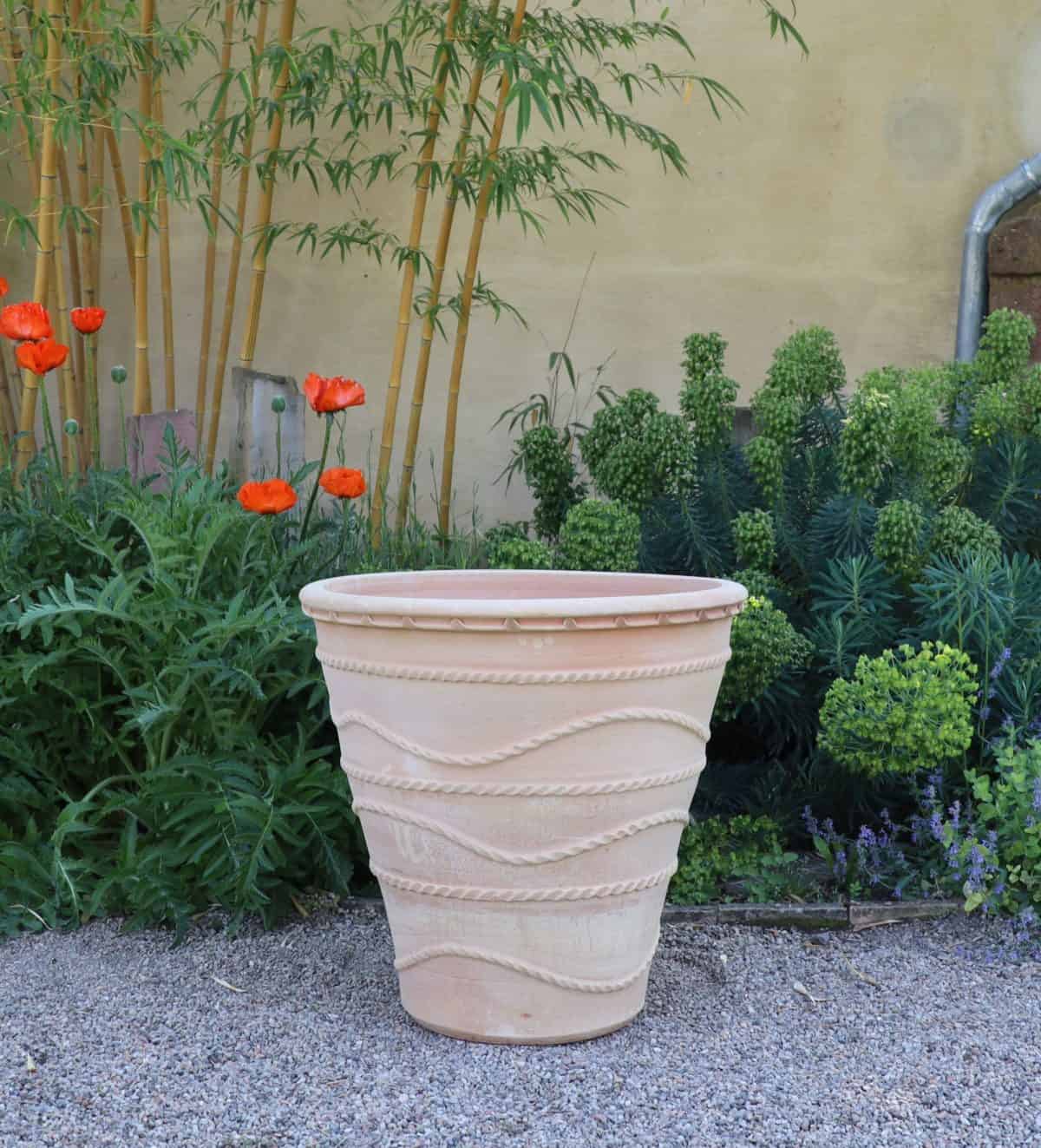 Ein winterfester großer Pflanzkübel „Acanthus“ mit einer Höhe von 60 cm und einem strukturierten, gewellten Design steht auf einem Kiesweg in einem Garten. Um den Topf herum stehen große grüne Acanthus-Pflanzen und blühende rote Blumen vor einer beigen Wand.