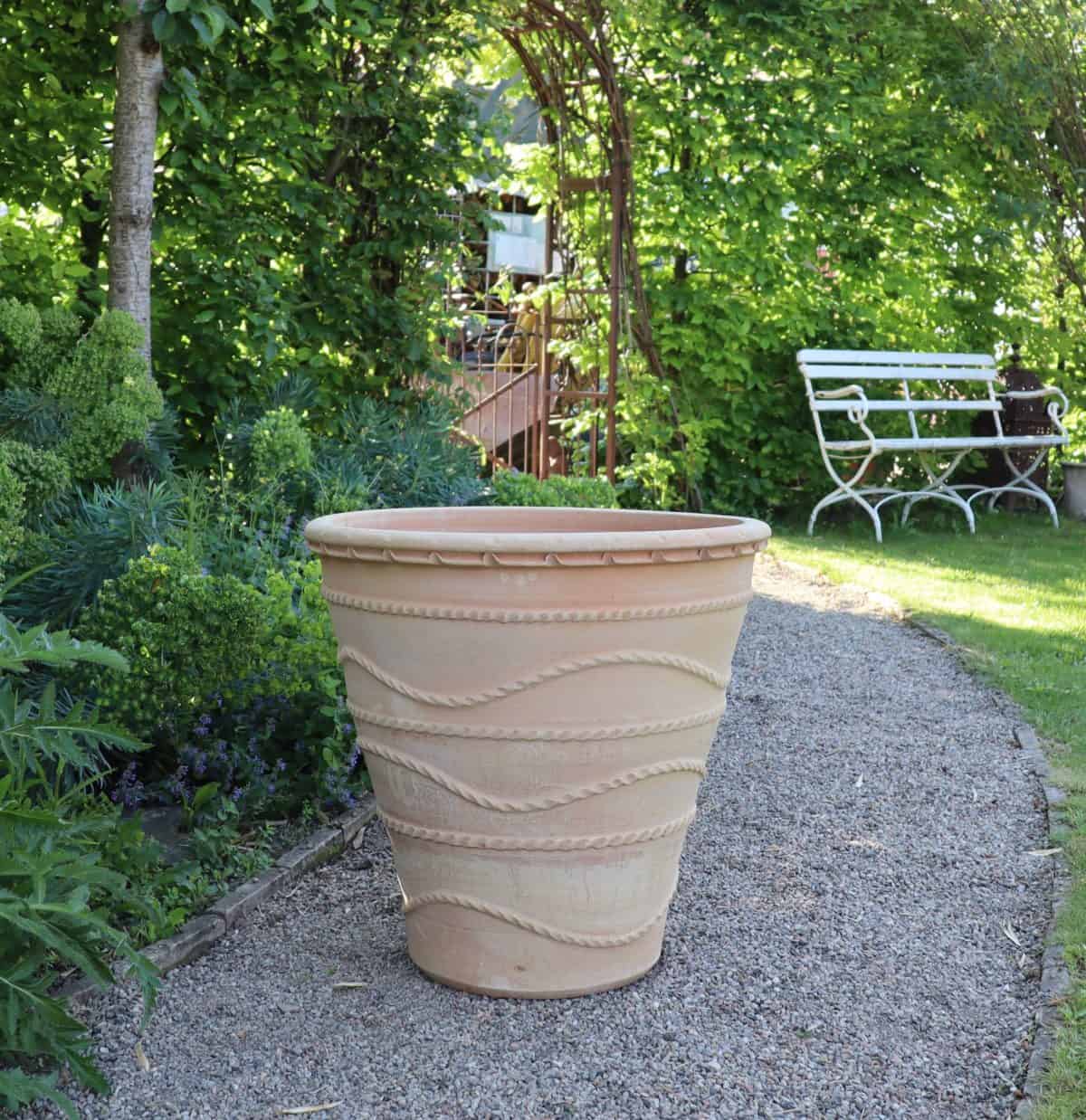 Ein großer, kunstvoll verzierter winterfester Pflanzkübel „Acanthus“, H=60 cm aus Terrakotta steht auf einem Kiesweg in einem üppigen, grünen Garten. Rechts ist eine weiße Metallbank teilweise sichtbar. Der Garten ist mit verschiedenen Pflanzen und Bäumen, darunter Acanthus, gefüllt und im Hintergrund ist ein Spalierbogen zu sehen.