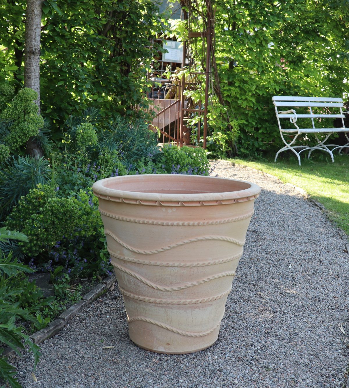 Der winterfester großer Pflanzkübel „Acanthus“, H=60 cm aus Terrakotta mit strukturiertem Acanthus-Muster steht auf einem Kiesweg in einem üppigen Garten. Um den Weg herum stehen verschiedene grüne Sträucher und Pflanzen. Im Hintergrund ist neben einem Holztor eine weiße Metallbank zu sehen.