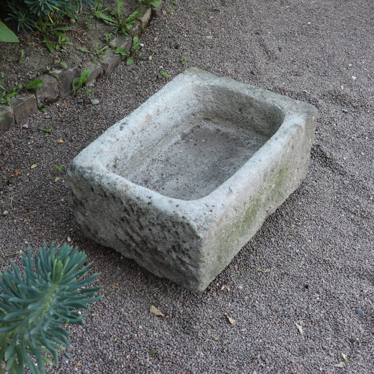 Ein nostalgischer Pflanzkübel aus Sandstein 70 x 50 x 30 cm steht auf einer Kiesfläche, umgeben von Grün. Der Kübel wirkt abgenutzt und verwittert, was darauf schließen lässt, dass er alt oder historisch sein könnte. Der Boden hat kleine Kieselsteine und am Rand ein paar Pflanzen.