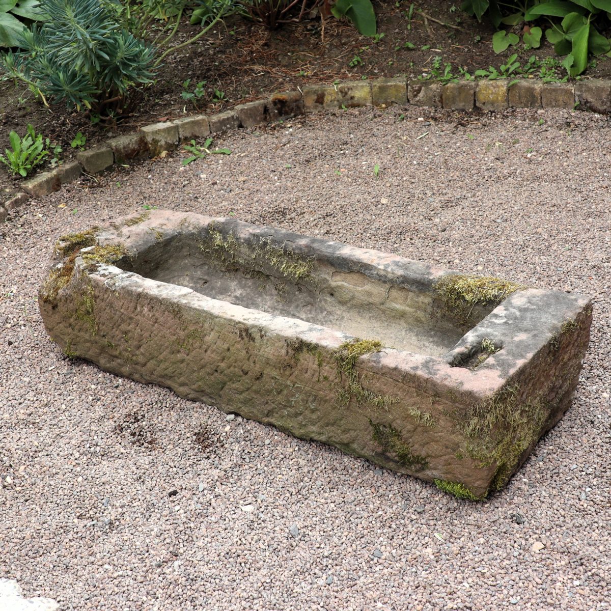 In einem Garten steht ein altertümlicher Sandsteintrog 118 x 40 x 24 mit rechteckiger Form auf Kies. Moos und Flechten wachsen auf seiner Oberfläche und verstärken sein antikes Aussehen. Der Trog ist von Pflanzen umgeben und von einer geschwungenen Linie kleiner Ziegel eingefasst.