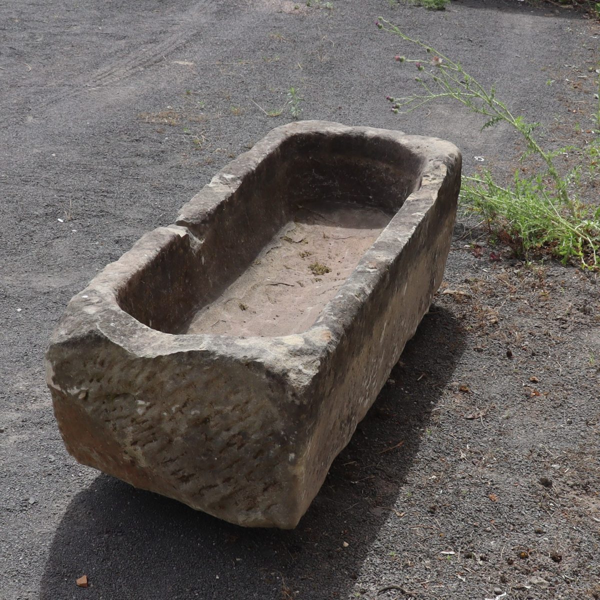 Ein verwittertes antikes Sandsteinbecken 154 x 50 x 36 steht auf einer Kiesfläche mit etwas überwuchertem Unkraut in der Nähe. Der Trog hat raue, unebene Kanten und einen großen Riss auf einer Seite, was auf Alters- und Abnutzungsspuren hinweist.