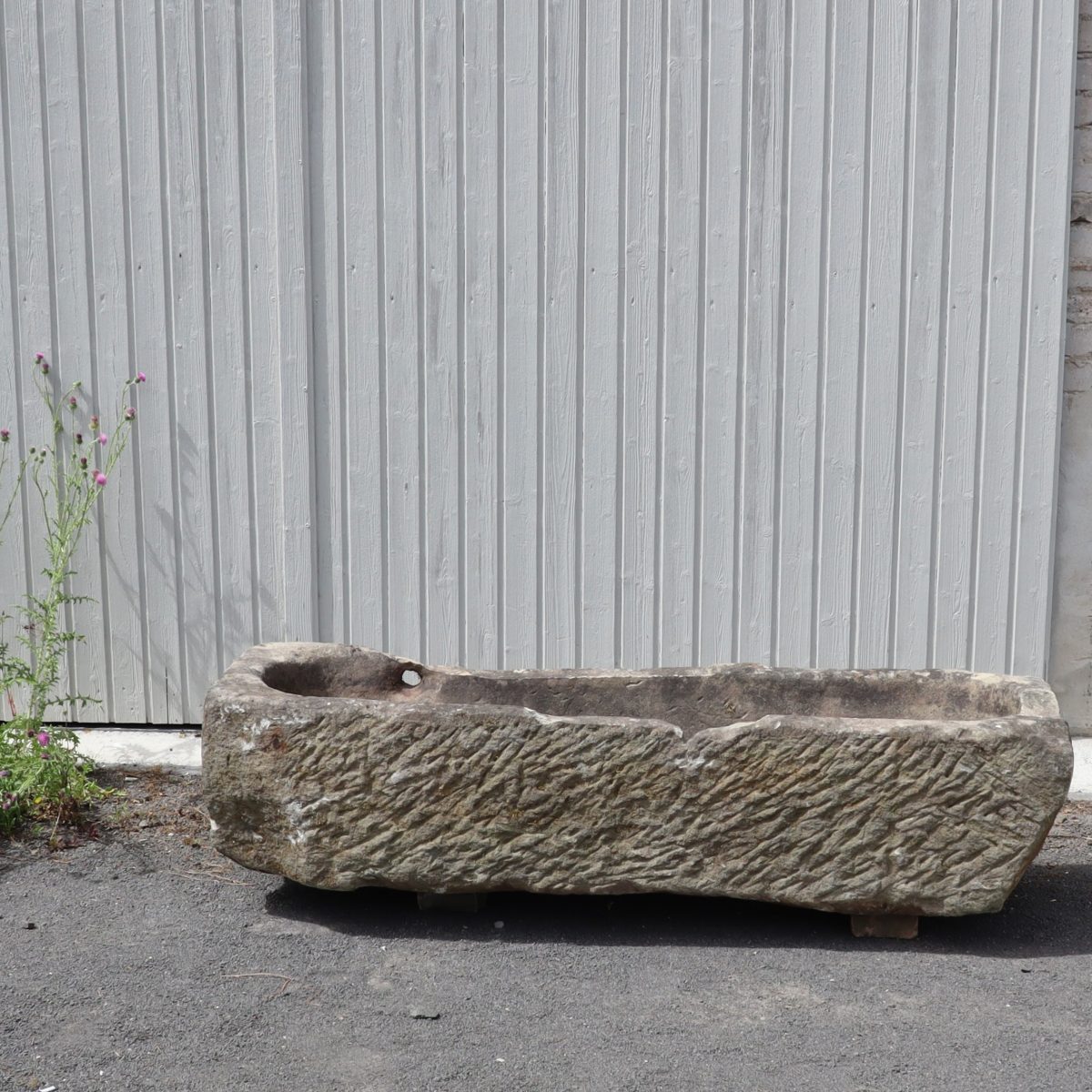 Ein verwittertes, rechteckiges antikes Sandsteinbecken (154 x 50 x 36) steht auf einer gepflasterten Fläche vor einer gewellten Metallwand. Der Trog weist altersbedingte Strukturen auf und in der Nähe wachsen ein paar Pflanzen.