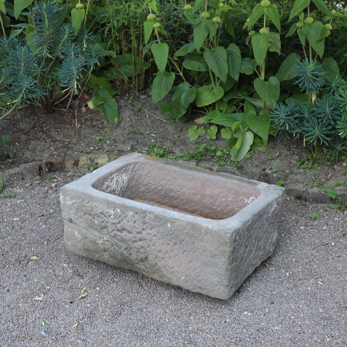 Ein altertümliches Sandsteinbecken 78 x 53 x 30 cm steht auf einer Kiesfläche vor üppigem grünem Laub. Der Trog wirkt verwittert und hat eine raue Textur, was ihm einen rustikalen Charme verleiht.