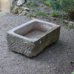 Ein altertümliches Sandsteinbecken mit den Maßen 78 x 53 x 30 cm und verwitterten, rauen Kanten steht auf einer Kiesfläche. Um das Becken herum erinnern einige Grünpflanzen und felsiges Gelände an einen Garten im Freien oder eine ländliche Umgebung.